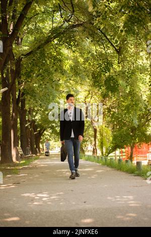 Giovane bel bussinessman che cammina lungo la strada nel parco e si perde nei suoi pensieri Foto Stock