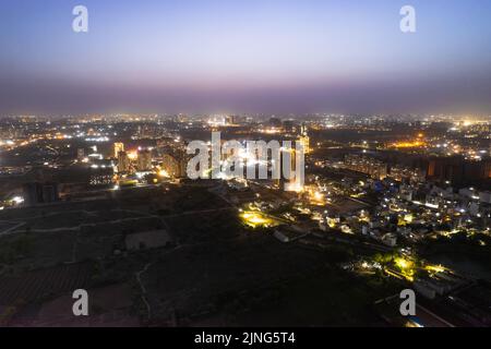 crepuscolo con droni aerei che mostra luci arancioni di strade, case e mercati che circondano un grattacielo con il paesaggio della città che si estende nel Foto Stock