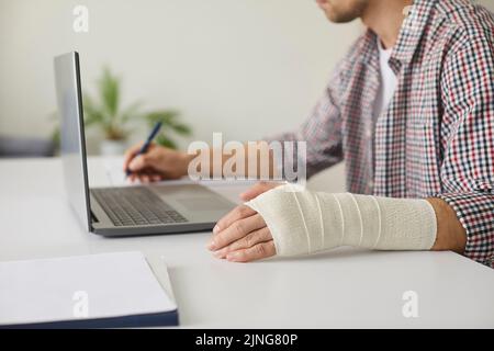 Uomo con fasciatura elastica avvolto intorno al polso seduto alla scrivania e al lavoro sul computer portatile Foto Stock