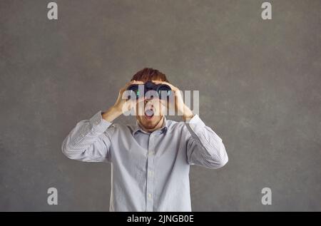 Uomo divertente che guarda a bocca aperta in binocolo in piedi isolato su sfondo grigio Foto Stock