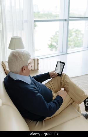Uomo anziano seduto sul divano e leggendo un e-book Foto Stock