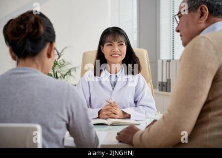 Ritratto in vita di un grazioso medico asiatico che indossa un cappotto bianco ascoltando i suoi pazienti anziani mentre si è consultati in un ufficio moderno Foto Stock