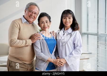 Ritratto di gruppo di un medico asiatico altamente professionale e di una coppia anziana affettuosa in piedi presso un ufficio moderno e guardando la macchina fotografica con sorrisi ampi Foto Stock