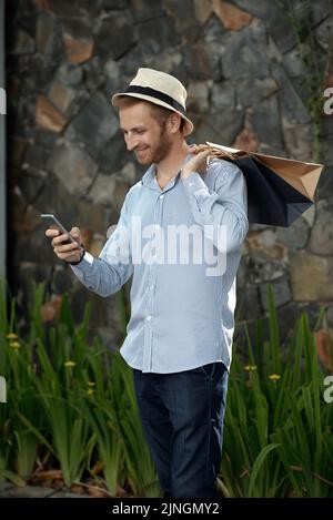 Bel giovane uomo che SMS sullo smartphone e tiene le borse della spesa all'aperto Foto Stock