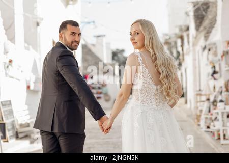 Ritratto di coppia che cammina, guardando indietro. Felice sposa indossando un lungo abito bianco con le mani che si interbloccano con lo spazio in tuta. Foto Stock