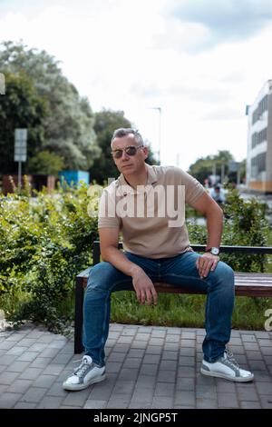 Ritratto di brutale uomo anziano in occhiali da sole seduti sulla panchina in cortile casa paesaggistico e riposante nelle giornate di sole tra le piante verdi. Stile di vita urbano Foto Stock