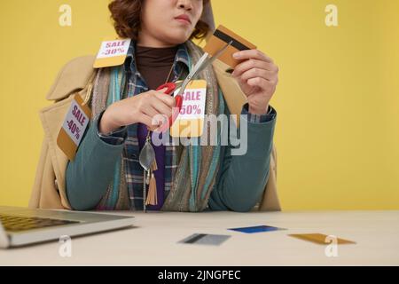 Donna asiatica che indossa strati di vestiti con le etichette di prezzo di vendita tagliando la sua carta di credito con le forbici Foto Stock