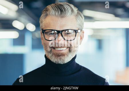 Primo piano ritratto fotografico di successo e felice uomo d'affari senior dai capelli grigi, investitore maschile con occhiali e barba guardando la fotocamera e sorridendo, capo che lavora in ufficio moderno Foto Stock