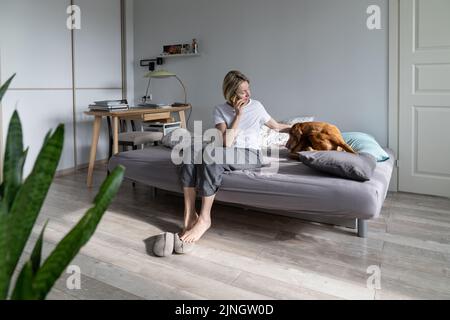 Donna bionda di mezza età parla al telefono con un amico e Strokes preferito cane a letto Foto Stock