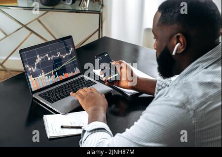 Grafici di trading finanziario sugli schermi di laptop e smartphone. Un commerciante afro-americano di successo, mediatore azionario, analizza le dinamiche di crescita nel mercato cripto, studia i rischi e le prospettive Foto Stock