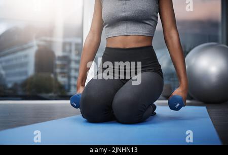 La resilienza è la superpotenza più grande. Una giovane donna irriconoscibile in ginocchio che tiene manubri mentre si esercita sul suo tappetino da palestra a casa. Foto Stock