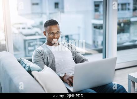 Rimani connesso anche nei weekend. Un bel giovane uomo che usa il computer portatile seduto su un divano a casa. Foto Stock