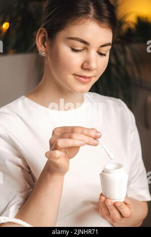 Ritratto di un estetista che raccoglie la crema con una spatola da una giara, pronta ad applicarla sul viso della donna. Test e pubblicità di prodotti, cura Foto Stock