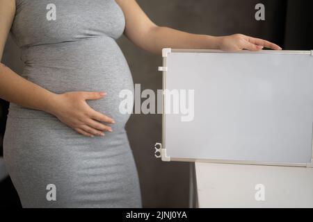 Giovane donna incinta con lavagna bianca. Donna in attesa felice tenendo vuoto foglio pubblicitario e toccando il suo ventre, sfondo grigio studio Foto Stock