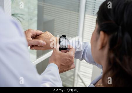Vista dall'alto del terapeuta amichevole che mostra al suo paziente anziano come controllare la frequenza cardiaca con l'aiuto dello smartwatch, primo piano Foto Stock