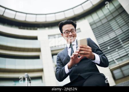 Uomo d'affari asiatico sorridente in occhiali che SMS sullo smartphone in aeroporto Foto Stock