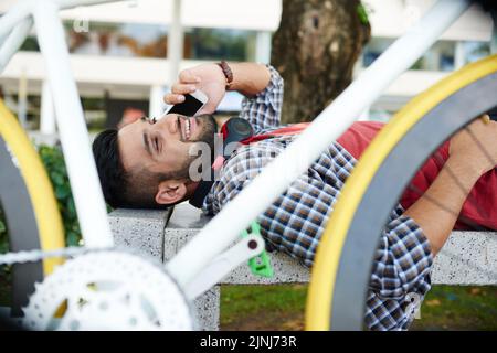 Bel ciclista indiano indossando una T-shirt rossa e una camicia a scacchiè sdraiata sulla panchina del parco pubblico verde e parlando con un amico su smartphone, ritratto girato Foto Stock