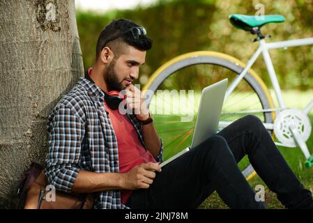 Bel giovane studente seduto sul prato del parco e utilizzando un moderno computer portatile mentre si prepara per l'esame universitario, bicicletta moderna in piedi dietro di lui Foto Stock
