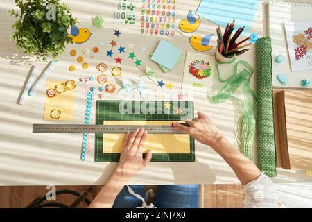Direttamente sopra la vista di una donna irriconoscibile utilizzando righello e coltello da taglio mentre si fa biglietto d'auguri da foglio di cartone, oggetti decorativi e strumenti sdraiati sulla superficie del tavolo Foto Stock