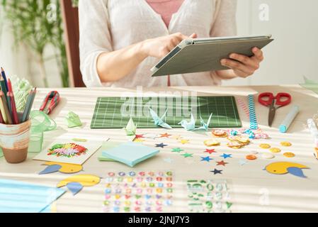 Giovane donna seduta al tavolo di legno e utilizzando un tablet digitale mentre si prende una pausa dalla produzione di gru origami, oggetti decorativi e strumenti in primo piano Foto Stock