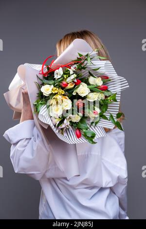 La donna bionda si alza con la schiena alla macchina fotografica e tiene sulle spalle un bouquet di rose, tulipani e verde sullo sfondo di una parete grigia. Con Foto Stock