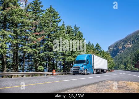 Semirimorchio industriale blu con cabina a tetto alto per il trasporto di carichi commerciali in semirimorchio posteriore che guida su una strada eccezionale lungo t Foto Stock