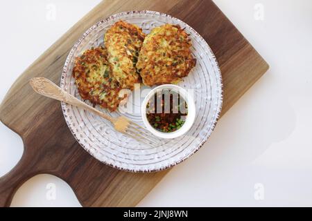 frittelle di zucchine vegetariane in un piatto bianco e salsa asiatica con spezie in una piccola ciotola bianca su un tagliere di legno. Sfondo bianco. Vegetaria Foto Stock