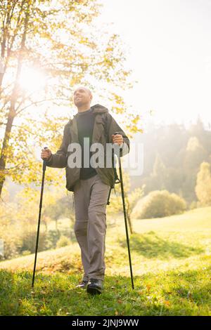 Sfondo naturale. Vista dal basso di un giovane viaggiatore maschio con bastoni da trekking in mano in piedi al sole. L'uomo sorridente gode della bellezza di n Foto Stock