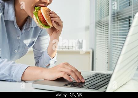 L'imprenditore femminile che mangia hamburger di pollo quando lavora su un computer portatile in ufficio Foto Stock