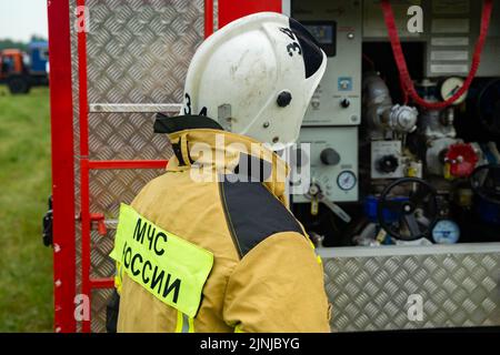 Un vigile del fuoco in un casco è in piedi vicino a un camion dei pompieri. Attrezzatura per camion antincendio. L'iscrizione sull'uniforme di EMERCOM della Russia Foto Stock