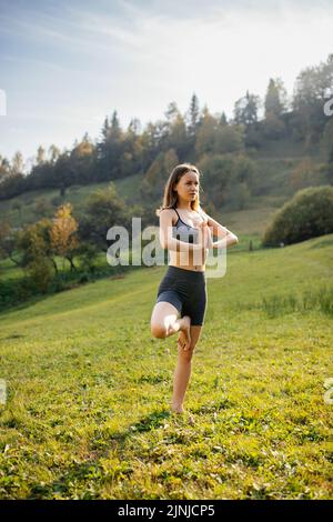 Yoga nel parco. Giovane donna atletica serena e pacifica si alza su una gamba e pratica il bilanciamento yoga all'aperto in una calda giornata autunnale. Concetto di a. Foto Stock