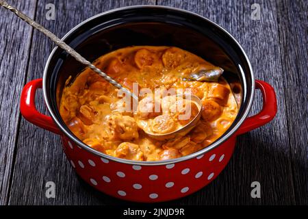 nakkikastike, salumi finlandesi in salsa di pomodoro cremosa in pentola rossa su tavolo di legno scuro con mestolo, primo piano Foto Stock