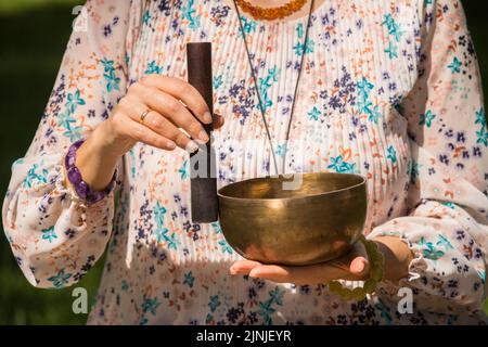 Dettaglio delle mani di una donna che tiene e suona una ciotola di canto tibetano durante una sessione di meditazione e terapia musicale in un parco. Foto Stock