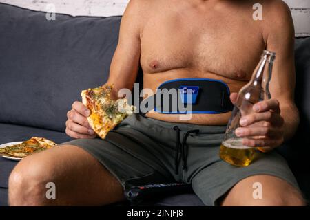 l'uomo seduto sul divano mangia pizza e beve birra mentre usa la cintura addominali dimagranti elettronica. stile di vita malsano Foto Stock