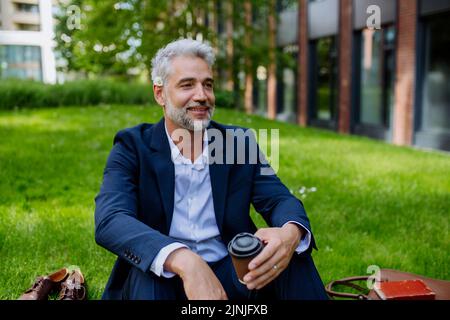 Uomo d'affari maturo che riposa e si siede a piedi nudi nel parco con una tazza di caffè, sentirsi libero, fuggire dal lavoro, concetto di equilibrio della vita lavorativa. Foto Stock
