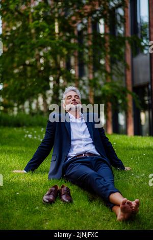 Uomo d'affari maturo che riposa e siede a piedi nudi nel parco, sentendosi libero, fuggendo dal lavoro, concetto di equilibrio di vita di lavoro. Foto Stock