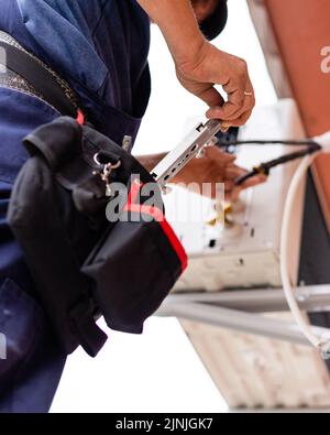 Un tecnico maschio collega l'unità di aria condizionata esterna alla parete del vecchio edificio di appartamenti. Primo piano dell'operatore che tira l'attrezzo fuori dalla sua borsa in vita. Selezionare Foto Stock