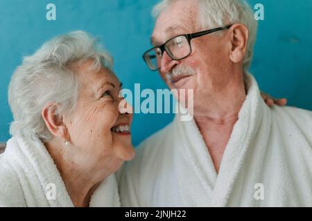 Ritratto di felice coppia anziana seduta insieme in accappatoio sul divano, avendo bel tempo a casa. Foto Stock