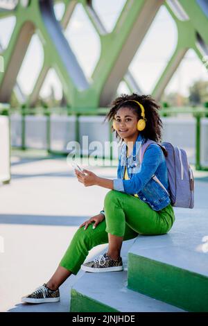 Ragazza adolescente multirazziale con zaino, cuffie e smartphone che riposa dopo la scuola nel ponte della città, ascoltando musica. Foto Stock