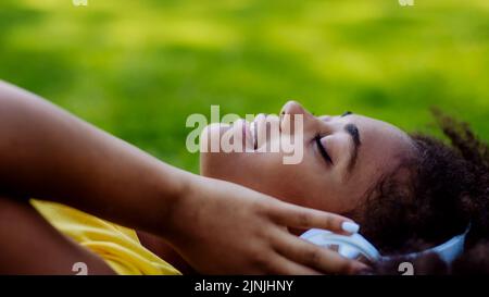 Ragazza multirazziale sdraiata in erba e godersi la musica in cuffie, vista laterale. Foto Stock