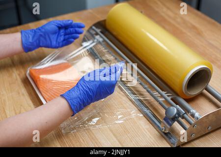 L'operaio in guanti monouso blu avvolge il pezzo di filetto di salmone sdraiato su un vassoio di plastica bianca in pellicola trasparente. Concetto di immagazzinaggio, mantenendo salato e. Foto Stock