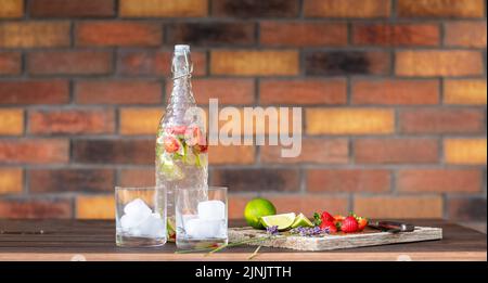 Bevanda rinfrescante con fragola e lime posta su un banco di legno. Due bicchieri con ghiaccio. Sfondo muro di mattoni. Foto Stock