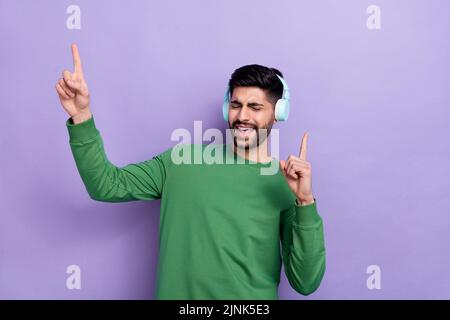 Foto di allegro bel ragazzo di buon umore indossare cuffie pullover verde occhi chiusi cantando danza isolato su sfondo di colore viola Foto Stock