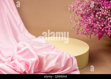 Podio geometrico cilindrico su sfondo beige, ricoperto da un tessuto di seta rosa. Sullo sfondo di piccoli fiori rosa. Podio per il prodotto Foto Stock