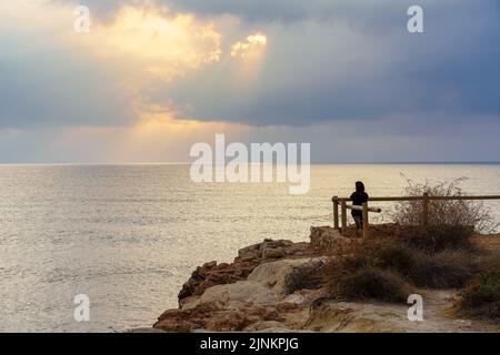 Donna che guarda all'orizzonte da un punto di vista al tramonto dorato sul mare. Foto Stock