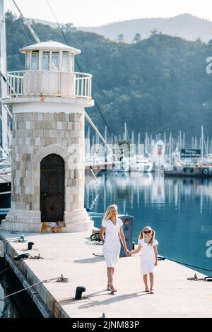 Felice madre millenaria e figlia bionda adolescente in abiti bianchi a piedi vicino al porto e porto turistico con yacht in una località turistica di mare con vela Foto Stock