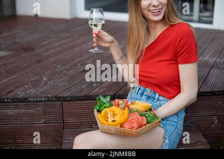 Ritratto di giovane gioiosa donna meravigliosa seduta su un gradino di veranda in legno, con bicchiere di vino bianco, verdure tagliate. Foto Stock