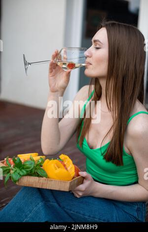 Ritratto di giovane felice donna impeccabile seduta sul gradino di veranda, bere vino dal vetro, tenere verdure tagliate. Foto Stock