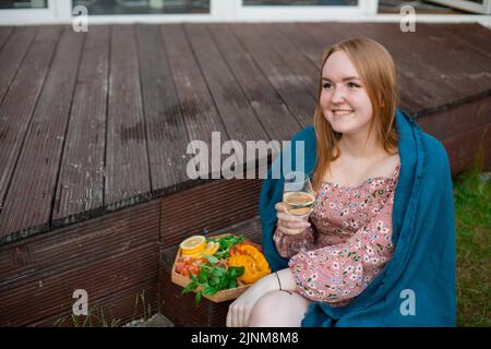 Giovane donna scuba sorridente seduta sul gradino di veranda vicino alla frutta, verdi su vassoio di legno, con bicchiere di vino bianco. Foto Stock