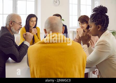 Gruppo multiculturale di persone sedute intorno al tavolo e che pregano insieme a Dio Foto Stock